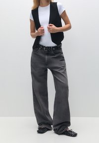 Person, der ein weißes kurzärmliges Oberteil, eine schwarze Weste, weit geschnittene verblasste schwarze Jeans mit Nieten-Gürtel und schwarze Ballettschuhe mit Nieten trägt.