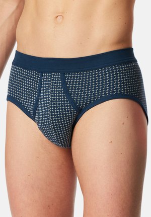 Mężczyzna w granatowych slipach z drobnym białym wzorem w kształcie krzyża, stojący, widoczny od tułowia do górnej części ud na jednolitym tle.