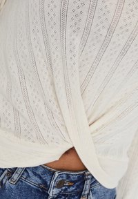 Top bianco lavorato a maglia con motivo a righe orizzontali e piccoli fori, annodato sul retro, abbinato a jeans blu a vita alta.