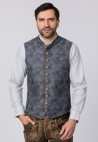 Dunkelblauer gemusterter Weste mit Stehkragen, Holzknöpfen und zwei Vordertaschen, über einem hellgrauen Langarmhemd getragen.