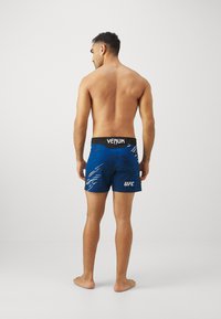 Mėlyni koviniai šortai su dinamišku raštu, juodas liemuo pažymėtas "VENUM" ir "UFC" logotipu, skirti sportiniam naudojimui, matomų kišenių nėra.