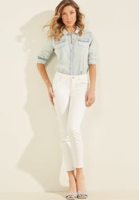 Cămașă din denim de un albastru deschis, cu dou�ă buzunare pe piept, asortată cu blugi albi, slim-fit, și tocuri argintii strălucitoare, pe un fundal neutru.