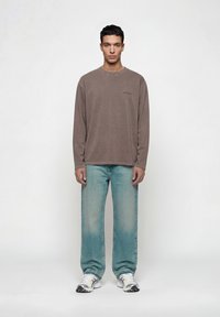 Άνδρας που στέκεται φορώντας καφέ μακρυμάνικη μπλούζα, ξεθωριασμένο μπλε τζιν και λευκά αθλητικά, μπροστά σε απλό φωτεινό φόντο.