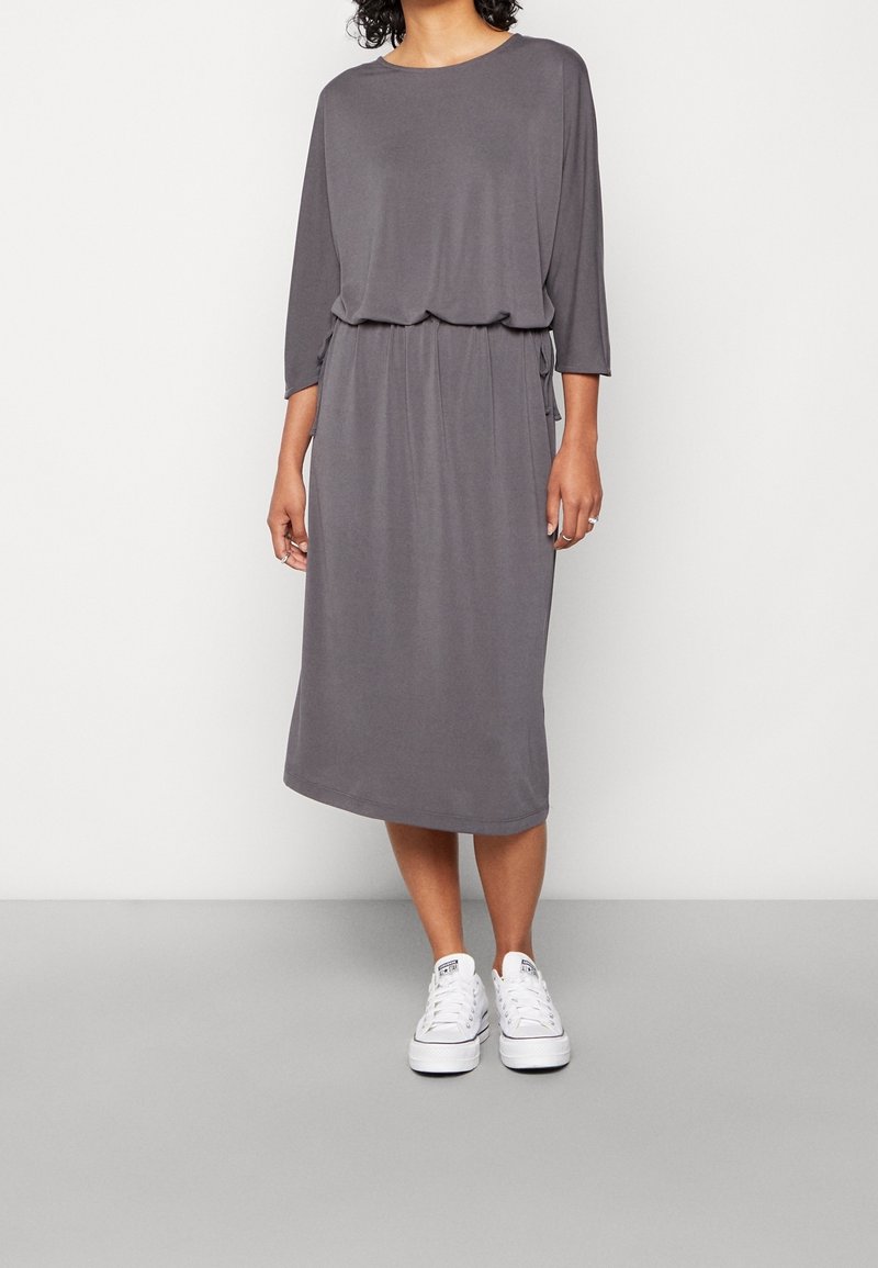 Femme portant une robe midi grise ample avec des manches trois-quarts et des baskets blanches, debout devant un fond clair uni.