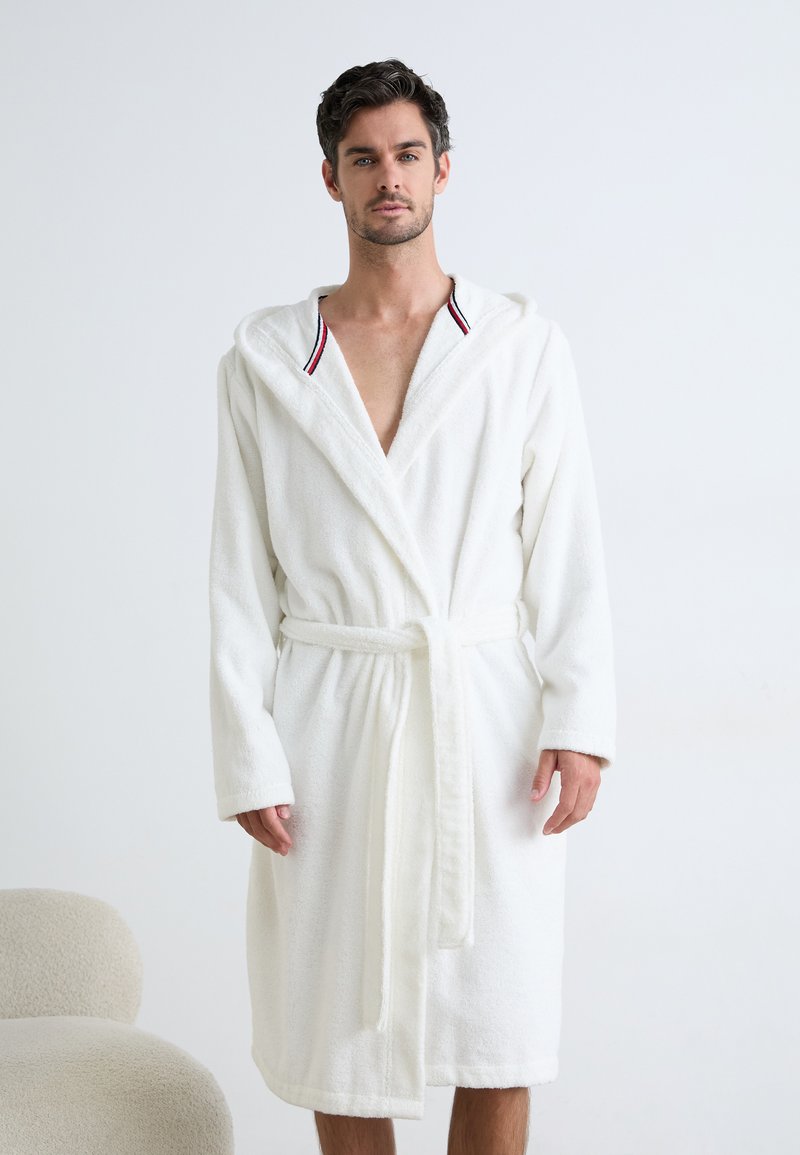 Accappatoio bianco fluffy con cappuccio, cinturino anteriore e dettagli a righe rosse e blu sul collo. Realizzato in morbido tessuto texturizzato.