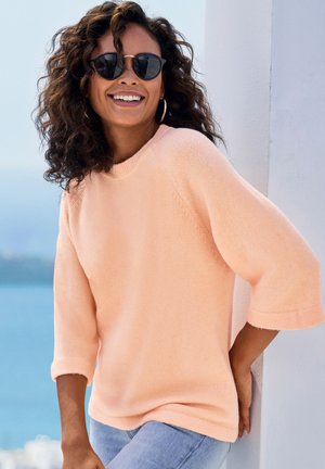 Femme aux cheveux bouclés et boucles d'oreilles créoles portant des lunettes de soleil, un pull pêche et un jean bleu, appuyée contre un mur blanc en plein air près de l'eau.