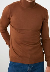 Maglione a collo alto marrone con una texture a coste e un design aderente, maniche lunghe e orlo aderente, abbinato a jeans blu.