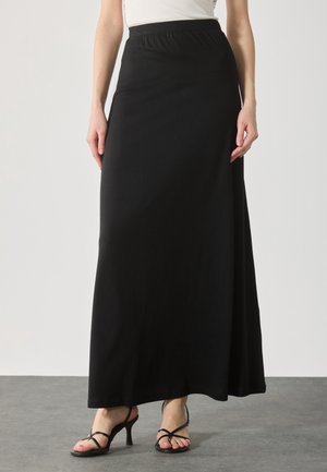 Femme portant une longue jupe noire à taille élastique et des sandales à talons noires à lanières, debout sur un sol gris.