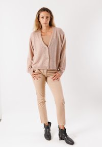 Cardigan rosa chiaro con chiusura a bottoni, tessuto testurizzato, vestibilità ampia. Indossato sopra un top beige, abbinato a pantaloni beige aderenti e stivali neri alla caviglia.