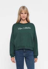 Blonde woman wearing dark green sweatshirt with "Gap Athletic" text, paired with blue jeans, standing against a plain white background.
