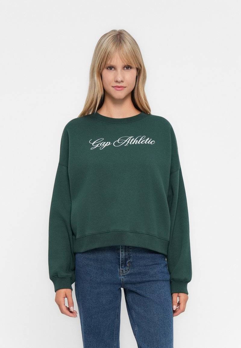 Blonde woman wearing dark green sweatshirt with "Gap Athletic" text, paired with blue jeans, standing against a plain white background.