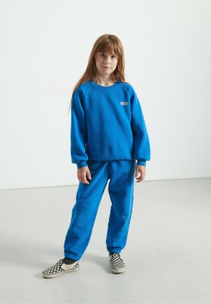 Sinine kapuutsiga sviiter ja sobivad püksid pehmest kangast. Sviitril on väike logo; pükstel on elastsed soonikud. Kingad on must-valge ruuduline.