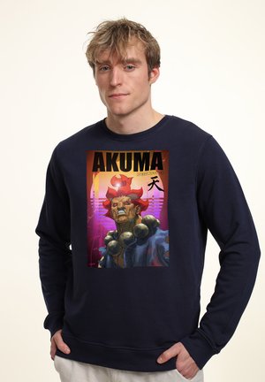 Jonge man die een marineblauwe trui draagt met een kleurrijke afbeelding van een woeste roodharige figuur en de tekst "AKUMA" op de voorkant.