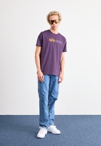 Violet medvilnės marškinėliai su "ALPHA INDUSTRIES" logotipu; šviesiai mėlynės džinsai; balti sneakeriai; rudi saulės akiniai. Paprastas dizainas ir laisvas pritaikymas.