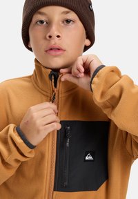 Veste en polaire marron avec une poche poitrine noire, détail de fermeture éclair et logo. Le col est haut et les manches sont dotées de poignets élastiques pour plus de chaleur.