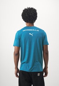 T-shirt turchese a maniche corte, con "MARSEILLE" e un logo Puma bianco stampato sulla parte alta della schiena. Texture morbida, vestibilità rilassata.