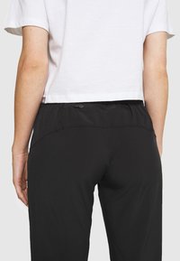 Pantalon noir léger avec une ceinture extensible dotée d'une poche zippée à l'arrière. Le tissu est lisse avec une finition mate.