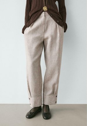 Persona che indossa pantaloni beige ampi con polsini abbottonati, un maglione marrone a coste, ballerine nere e una collana con ciondolo d'oro.