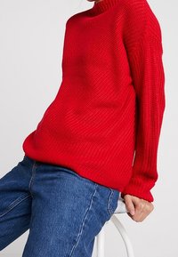 Röd stickad tröja med hög hals och texturmönster. Oversized passform med långa ärmar. Parat med blå denimjeans.