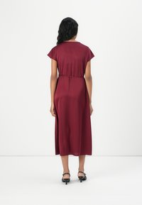 Robe bordeaux à manches courtes, avec une ceinture à la taille et une longueur au genou. Tissu lisse et fluide avec un éclat subtil. Talons noirs visibles en dessous.