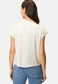 Hellbeige Bluse mit kurzen Ärmeln und hohem Kragen, glatter Textur und einem Schlüssellochverschluss im Rücken. Kombiniert mit blauen Jeans mit Gesäßtaschen.