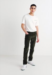 Vīriešu baltā kokvilnas T-kreklā ar zaļu logotipu, apvienots ar tumši zaļām korduroja biksēm. Viņš valkā baltas kedas ar zaļiem akcentiem.