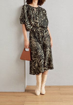 Femme portant une robe midi imprimé animal noir et beige, tenant un sac à main marron et portant des bottines beiges, debout à l'intérieur.