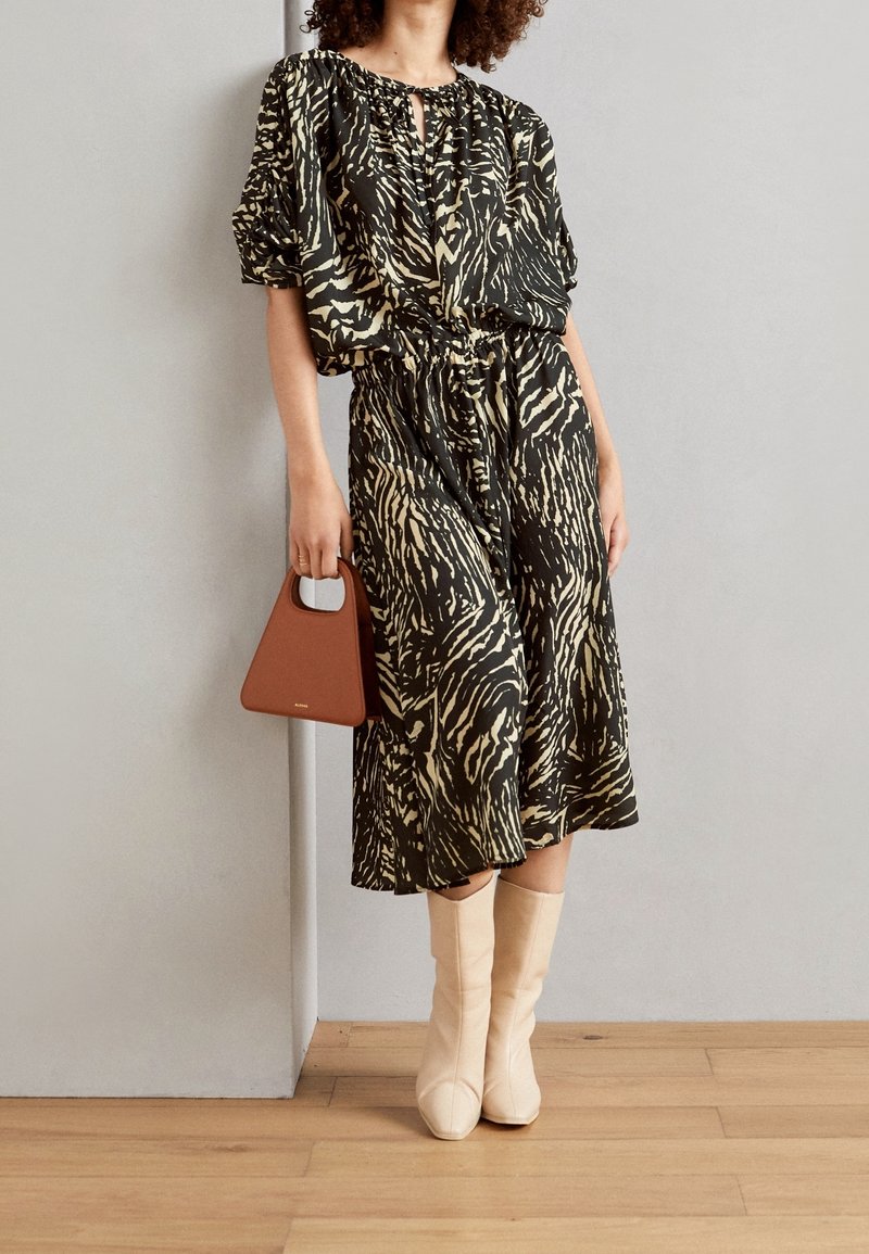 Femme portant une robe midi imprimé animal noir et beige, tenant un sac à main marron et portant des bottines beiges, debout à l'intérieur.