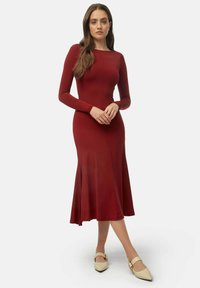 Robe longue ajustée à manches longues de couleur bordeaux avec un ourlet évasé. Sa texture lisse et ses coutures verticales mettent en valeur sa silhouette. Paire de chaussures à talons pointus beige.