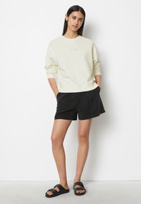 Felpa beige chiaro con spalle drop e polsini a coste, abbinata a pantaloni corti neri. Modella che indossa sandali neri, in piedi su uno sfondo bianco.