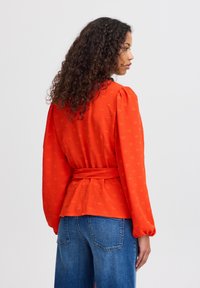 Blouse rouge texturée avec des manches bouffantes et une taille ceinturée, présentant un motif subtil. Associée à un jean en denim bleu taille haute.
