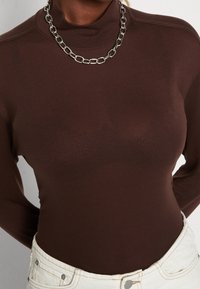 Brun långärmad polokrage med en mjuk textur och lätt lyster. Har ett silverfärgat kedjearmband och är matchad med vita byxor.