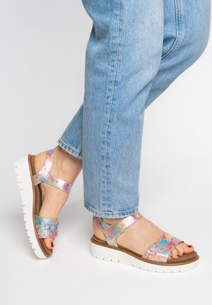 Feet in light blue jeans wearing colorful floral-patterned metallic sandals with white platform soles and red toenail polish.