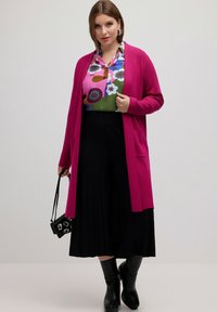 Fuchsia Cardigan, lange Ärmel, offenes Design, kombiniert mit einer blumigen Bluse in Pink und Grün sowie einem schwarzen plissierten Rock. Schwarze kniehoge Stiefel.