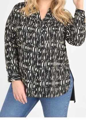 Mujer con blusa de manga larga con patrón abstracto en blanco y negro combinada con vaqueros azules, posando con una mano levantada y la otra apoyada en el muslo.