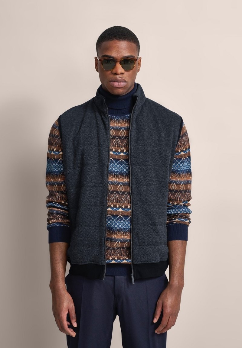 Gepolsterte, dunkelgraue Weste mit hohem Kragen über einem gemusterten braunen, blauen und beigefarbenen Strickpullover. Mit gerippten Bündchen und schwarzem Taillenbund.