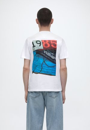 Personne aux cheveux courts porte un T-shirt blanc avec un graphisme coloré de 1985 et un jean bleu, tourné dos à la caméra sur un fond blanc.