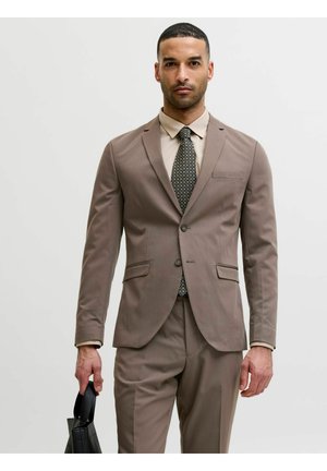 Hombre con traje color topo y corbata con estampado, sosteniendo un maletín de cuero negro sobre un fondo claro y liso.