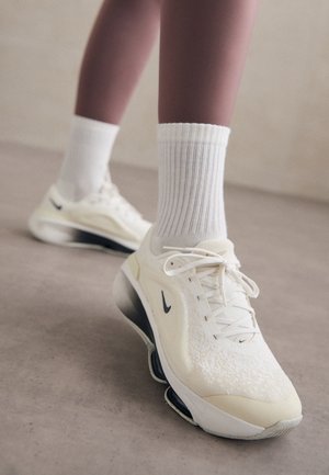 Osoba nosząca białe buty sportowe Nike z czarnym logo Swoosh i białe prążkowane skarpetki, stojąca na gładkiej podłodze.