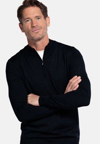 Donker marineblauwe pullover trui met een kwart rits en een hoge kraag. Gebreide textuur en geribbelde manchetten. Gedragen over een wit shirt.