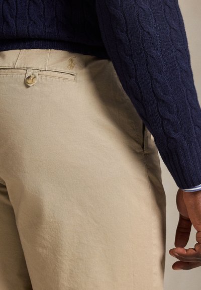 Άνδρας που φοράει μπεζ παντελόνι με πίσω τσεπάκι με κουμπί και σκούρο μπλε πλεκτό πουλόβερ με σχέδιο κοτσίδες, ορατό από τη μέση έως το χέρι από πίσω.