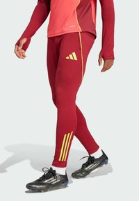 Rote sportliche Leggings mit gelben diagonalen Streifen und einem adidas-Logo, kombiniert mit schwarzen Fußballschuhen und weißen Knöchelsocken.