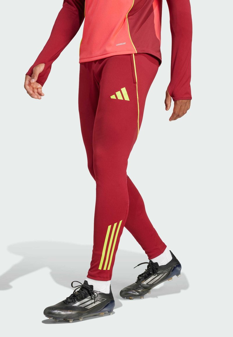 Rote sportliche Leggings mit gelben diagonalen Streifen und einem adidas-Logo, kombiniert mit schwarzen Fußballschuhen und weißen Knöchelsocken.