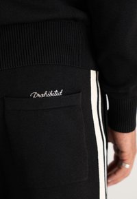 Black knit pants with white side stripes and a back pocket featuring white embroidered text "Prohibited" near a person's hand with rings.