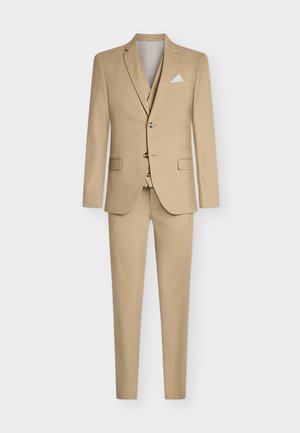 Beige kolmiosainen puku, jossa on säädettävä takki, jossa on kaksi nappia, viisto kaulus ja hienovarainen rakenne, yhdistelemässä räätälöidyt housut ja taskuliina.