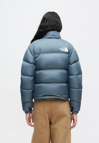 Osoba s dlhými tmavými vlasmi, oblečená v modrom púzdrovom kabáte a svetlohnedých nohaviciach, otočená chrbtom k jednému bielému pozadiu.