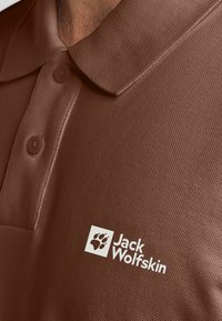 Polo shirt marrone con un tessuto texturizzato, dotato di colletto e due bottoni, e un logo bianco sul lato sinistro.