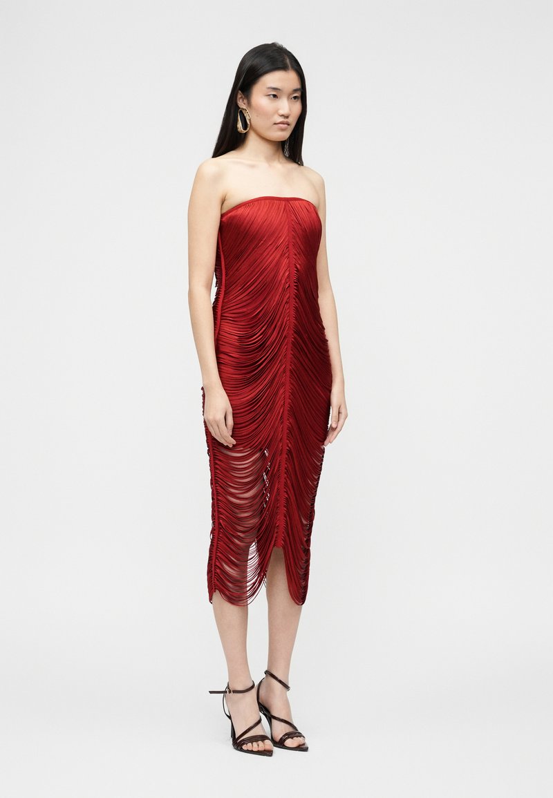 Femme portant une robe rouge sans bretelles, longueur genou, avec des franges superposées, et des sandales noires à talons hauts, debout devant un fond uni.