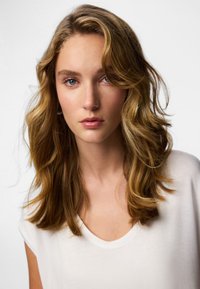 Hosszú, hullámos, világosbarna hajú modell fehér V-nyakú pólóban. Sima bőr, semleges smink és kék szemek, egyszínű háttér előtt.