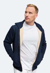 Uomo biondo che indossa un hoodie blu navy con rivestimento beige sopra una maglietta bianca, che guarda di lato con le mani in tasca su uno sfondo semplice.